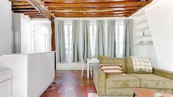Ein gemütliches Wohnzimmer mit einem grünen Sofa, Holzbalkendecke, einem weißen Teppich und einem Essbereich mit Tisch und Stühlen.