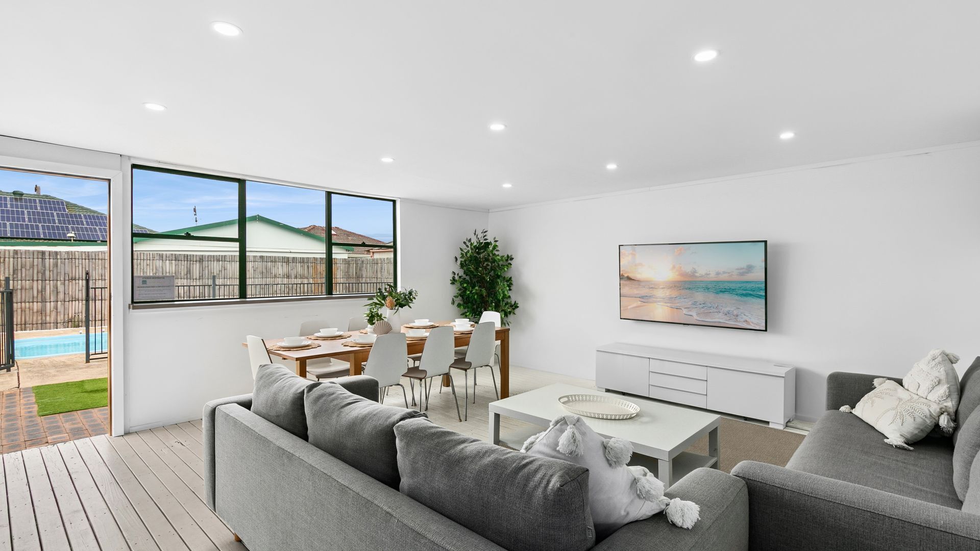 A modern living room with a pool view, a dining area, and a TV on the wall.