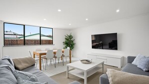 A modern living room with a dining area, a large window with a view, a flat-screen TV, and a grey sofa.