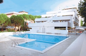 A rectangular swimming pool with a tiled deck, surrounded by a white building with balconies and a clear sky.