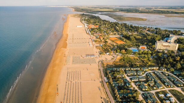 An aerial view of a coastal resort with a long sandy beach, multiple hotel buildings, and a marina. - Pool and beach at your doorstep (Bibione)