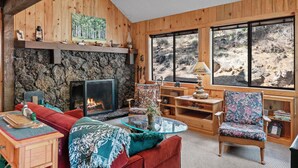 A cozy living room with a stone fireplace, wooden walls, a red sofa, a glass coffee table, and a large window with a view of a rocky landscape. - RR015 | Lovely Ranch cabin nestled in the woodsy Central Oregon landscape. (Black Butte Ranch)