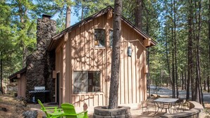 A wooden cabin with a stone chimney, a grill outside, and outdoor seating. - RR015 | Lovely Ranch cabin nestled in the woodsy Central Oregon landscape. (Black Butte Ranch)