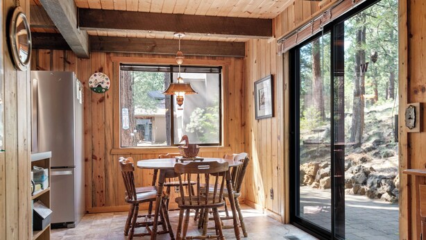 A wooden interior with a dining table and chairs, a refrigerator, and a large window offering a view of trees. - RR015 | Lovely Ranch cabin nestled in the woodsy Central Oregon landscape. (Black Butte Ranch)