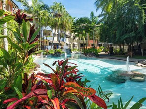Un espace piscine aménagé dans un complexe hôtelier, avec des palmiers, un élément décoratif aquatique et un bâtiment doté de balcons.