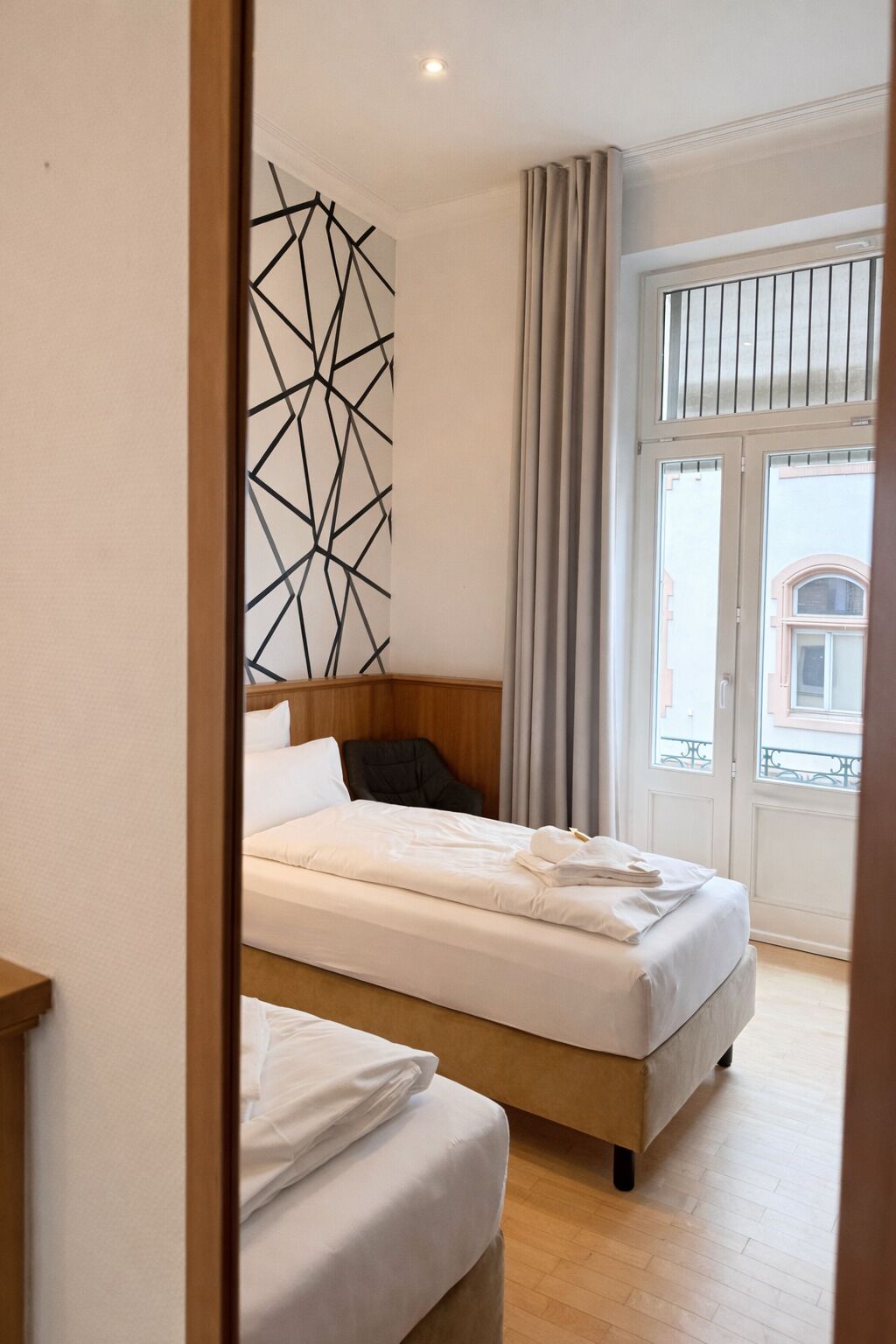Ein Hotelzimmer mit Bett, Holz-Kopfteil, Wanddekoration mit geometrischem Muster und Ausblick durch ein mit Vorhängen versehenes Fenster.