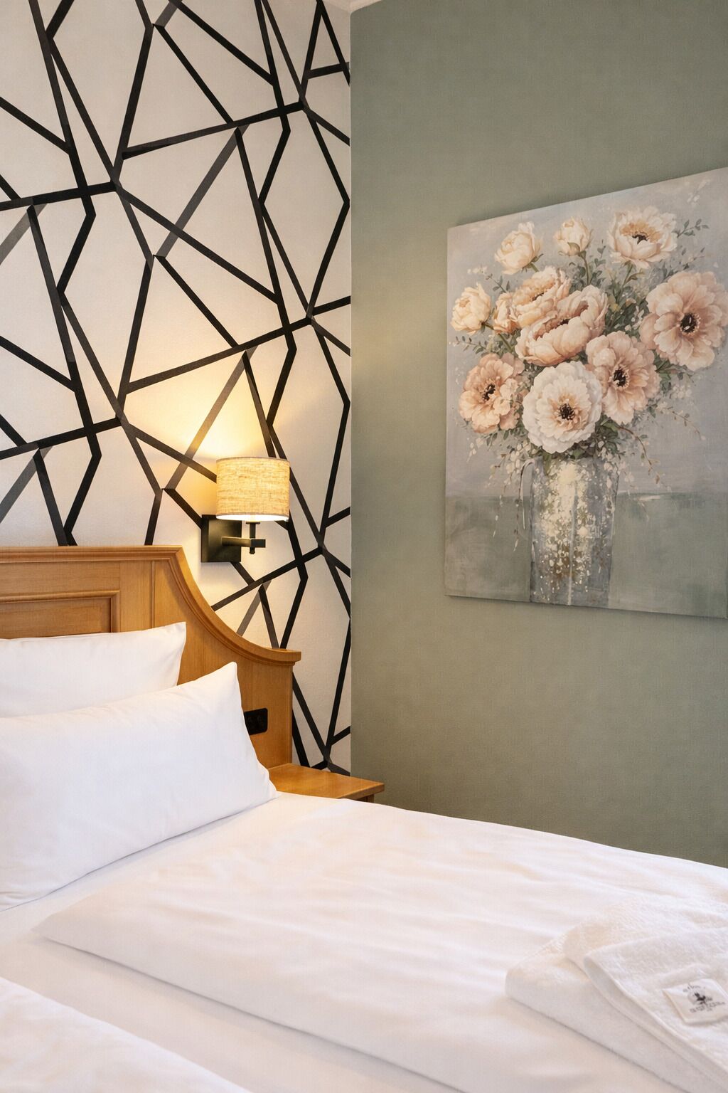 Une chambre d’hôtel avec un mur orné d’un motif géométrique, un tableau représentant des fleurs, un lit avec du linge de maison blanc et une lampe de chevet.