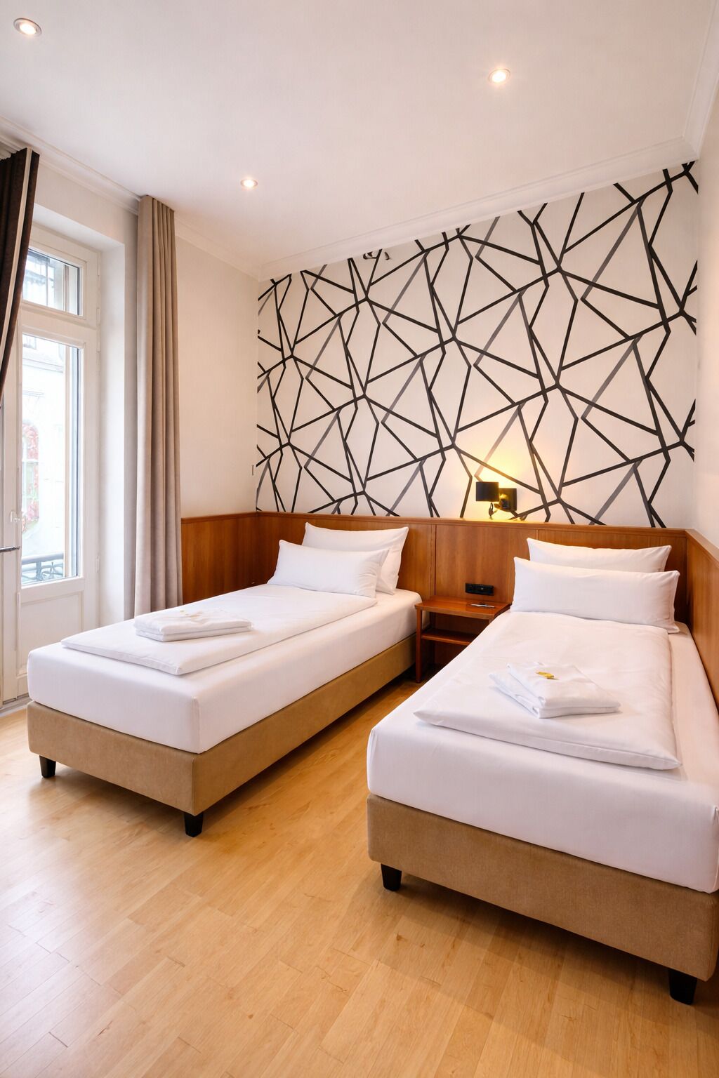 Ett hotellrum med två sängar, sänggavlar i trä, ett geometriskt mönster på väggen, en sänglampa och ett fönster med gardiner.