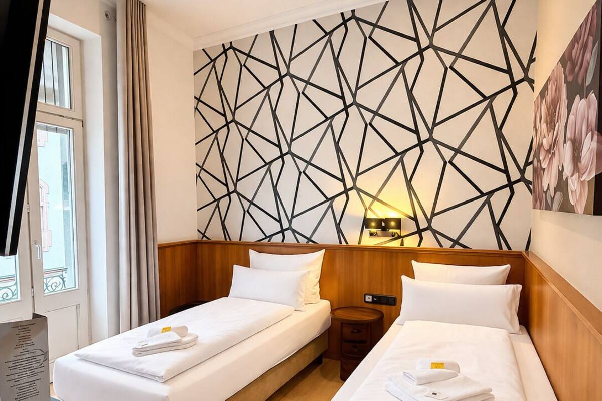 A hotel room with two single beds, a wooden headboard, a geometric wall design, a bedside table with a lamp, and a window with curtains.