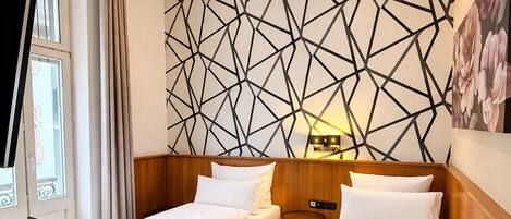 A hotel room with two single beds, a wooden headboard, a geometric wall design, a bedside table with a lamp, and a window with curtains.