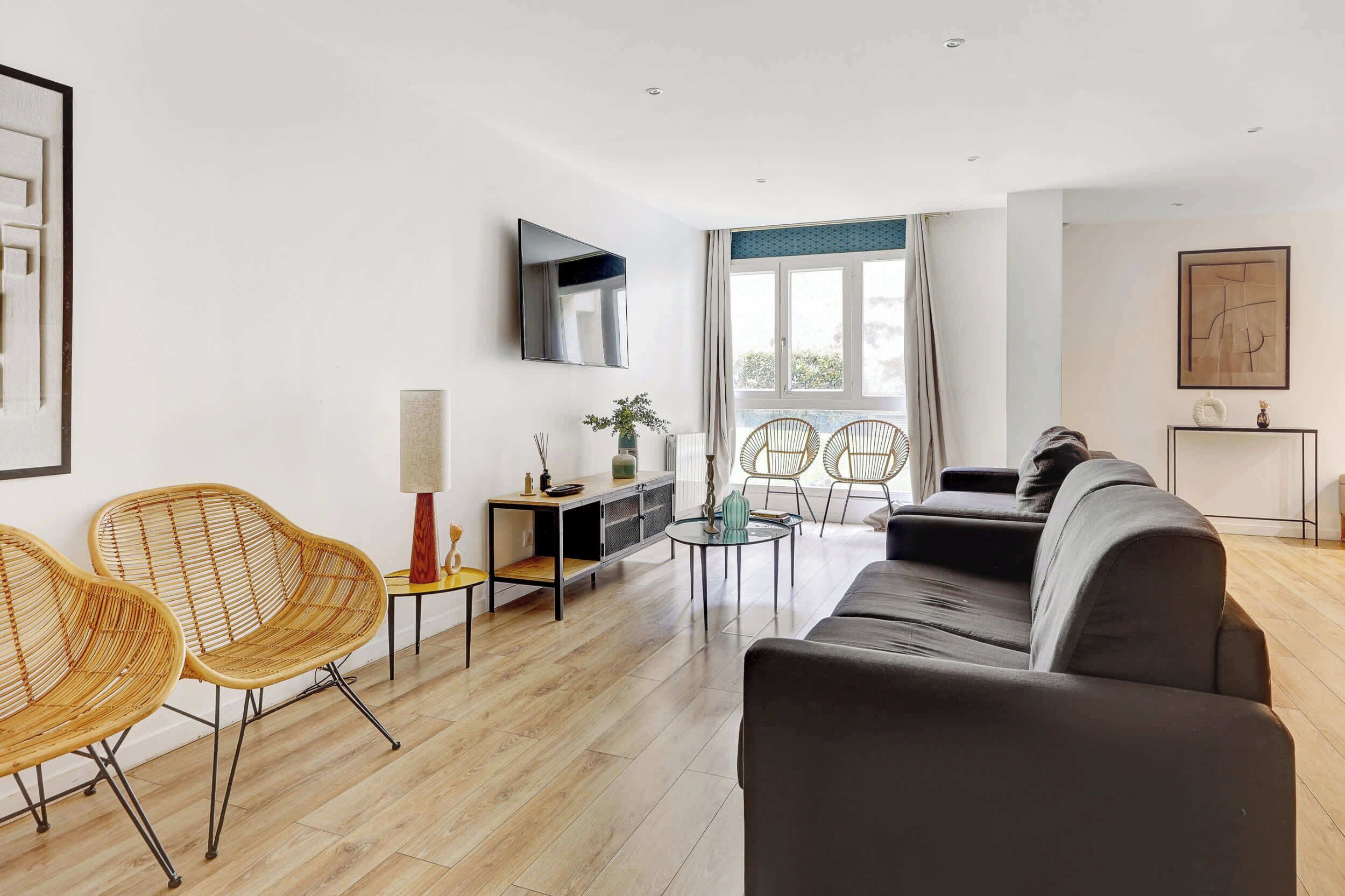A modern living room with a sofa, two wicker chairs, a small table, a TV mounted on the wall, and a large window with curtains.