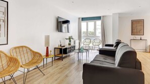 A modern living room with a sofa, two wicker chairs, a small table, a TV mounted on the wall, and a large window with curtains. - Spacious 4BDR in Buttes Chaumont pour 12 pers (Paris)