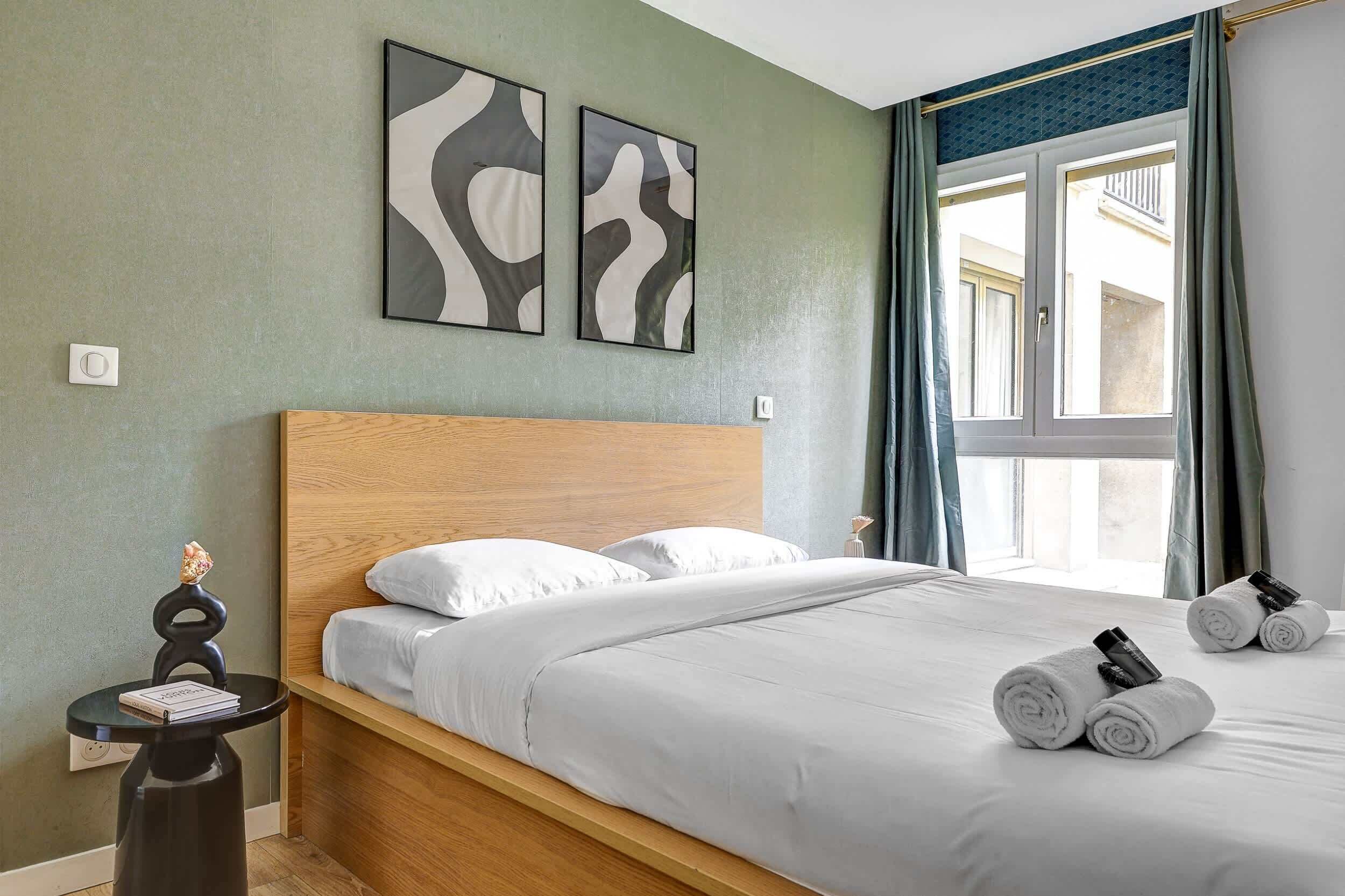 A neatly made bed with white linens and rolled towels, a wooden headboard, and two abstract wall art pieces.