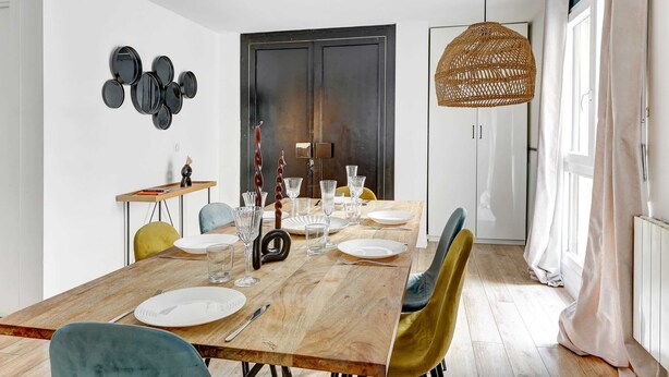 A dining area with a wooden table set for six, surrounded by chairs, and a wall-mounted art piece above the door. - Spacious 4BDR in Buttes Chaumont pour 12 pers (Paris)