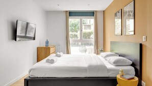 A modern bedroom with a bed, a TV mounted on the wall, a window with curtains, and a yellow side table. - Spacious 4BDR in Buttes Chaumont pour 12 pers (Paris)