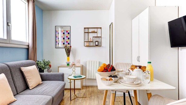 Ein modernes Wohnzimmer mit einem grauen Sofa, einem kleinen runden Tisch mit einer Vase sowie einem Frühstückstisch mit Früchten und einem Croissant.