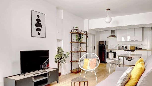 Una sala de estar moderna con un televisor de pantalla plana, un sofá con cojines, una mesita de madera y una silla blanca.