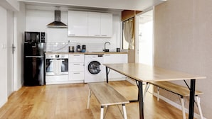 Una cocina compacta con armarios blancos, un refrigerador negro, un microondas, una estufa, una lavadora y un banco de madera.