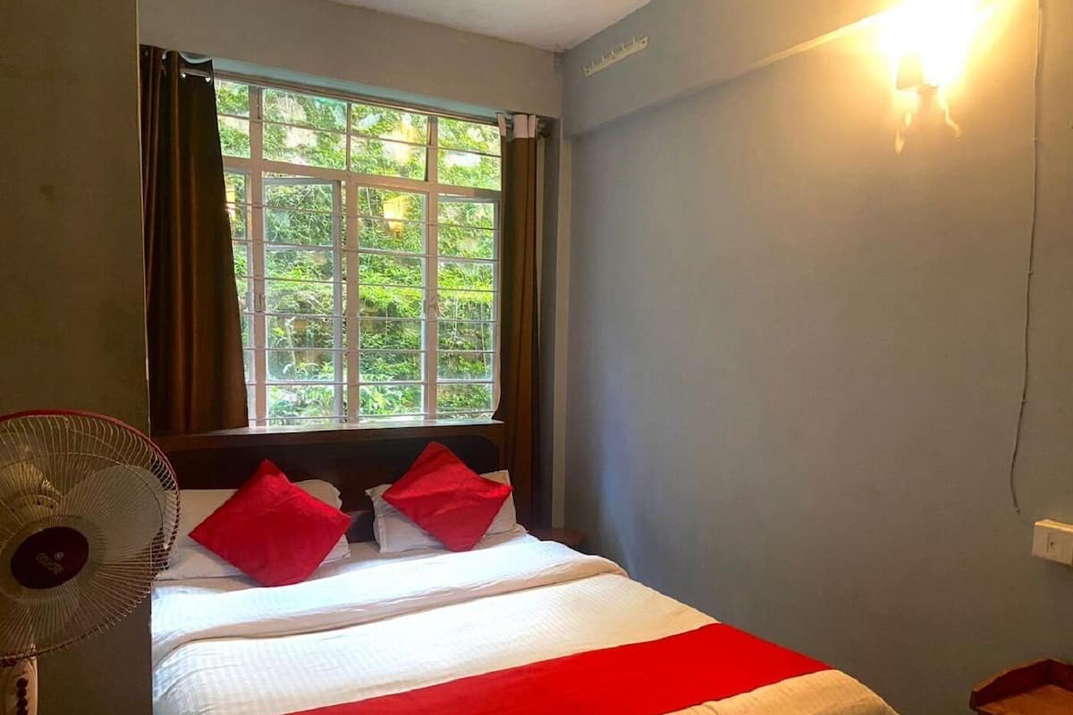 A bedroom with a bed, a window with curtains, a wall-mounted light, and a standing fan.