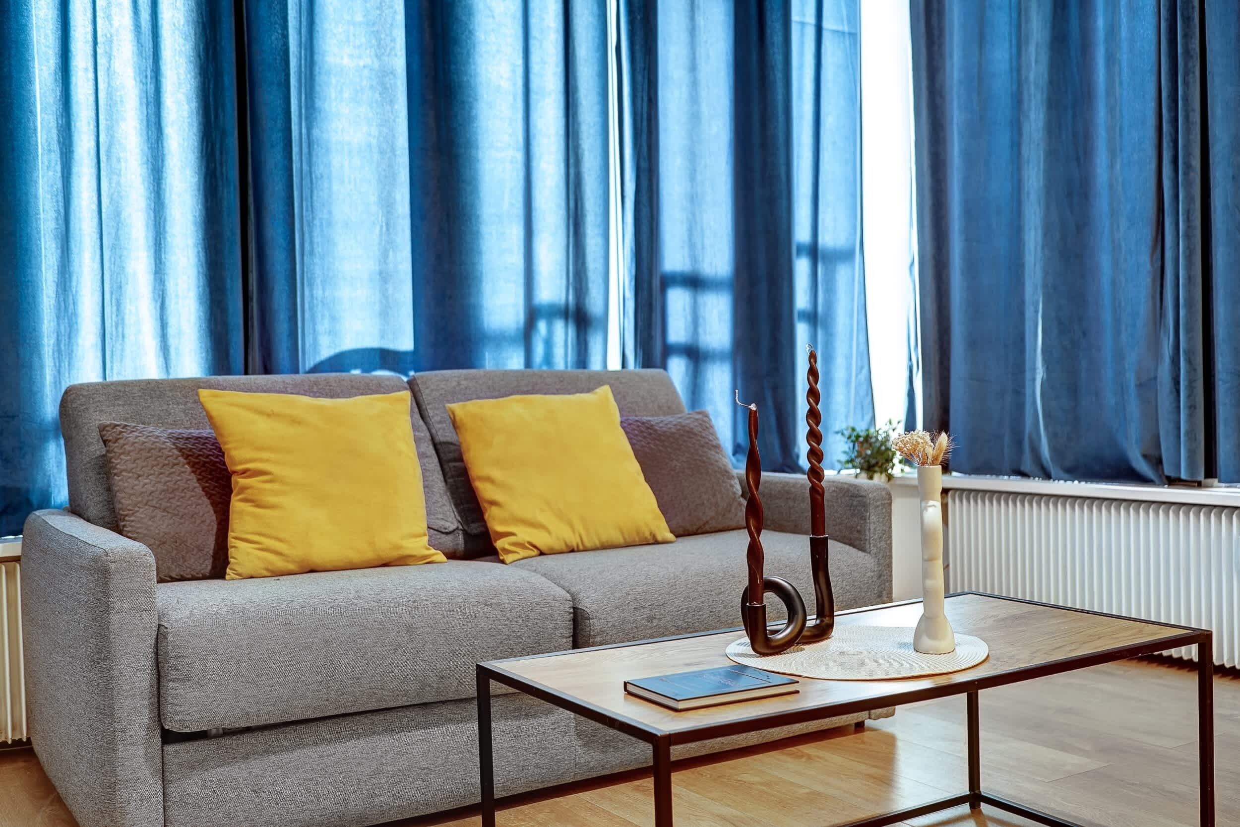 A living room with a grey sofa, yellow cushions, a wooden coffee table, and a vase with dried plants.