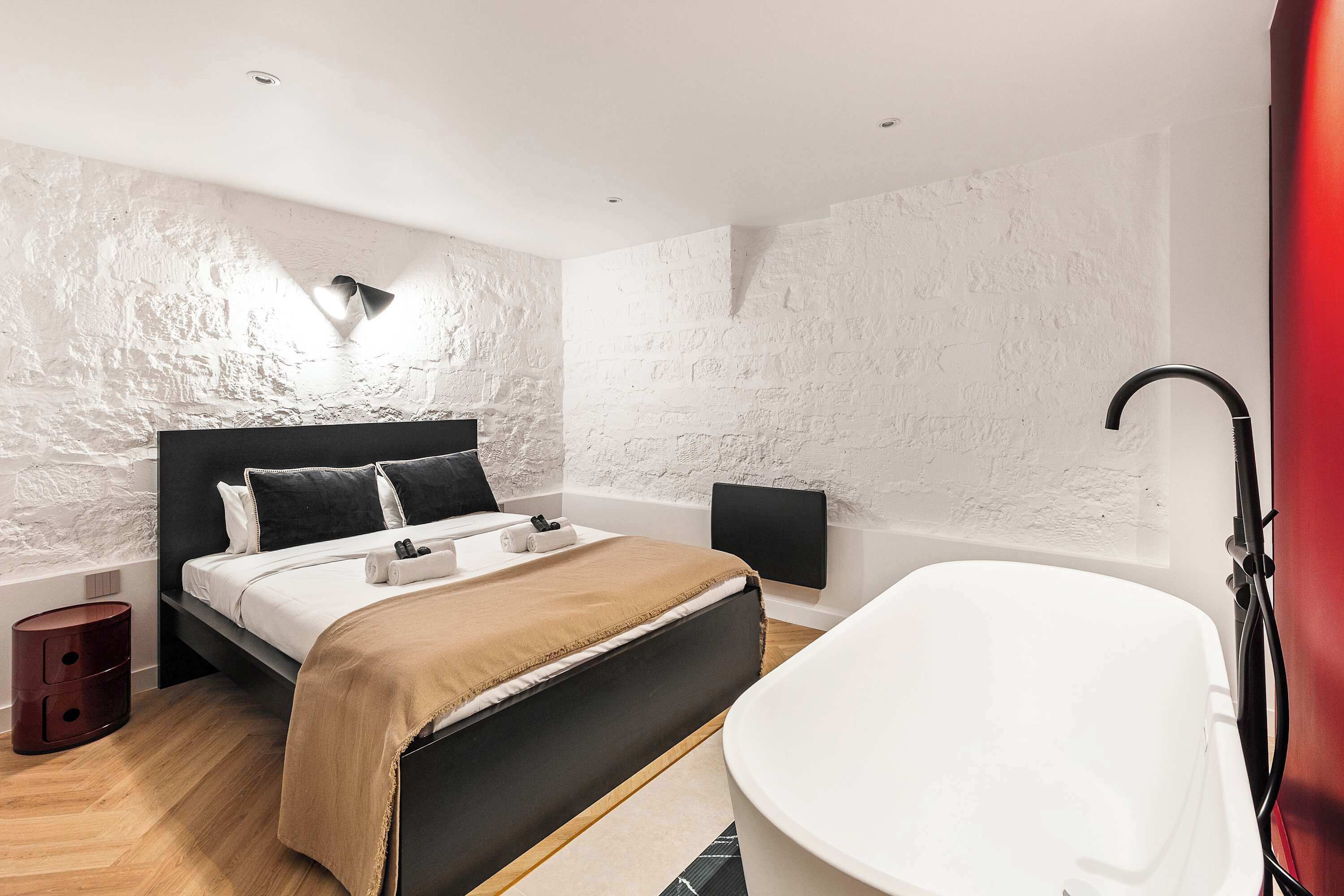 A modern bedroom with a large bed, a bedside table, a wall-mounted light, and a bathtub.