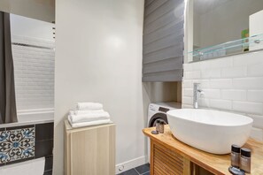 A modern bathroom with a large oval sink, a glass shower enclosure, a built-in washing machine, and a wooden vanity.