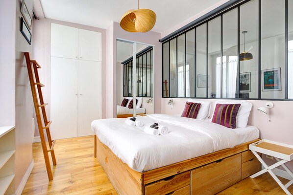 A bedroom with a wooden bed, white bedding, a ladder, a white desk, and a large window with blinds.