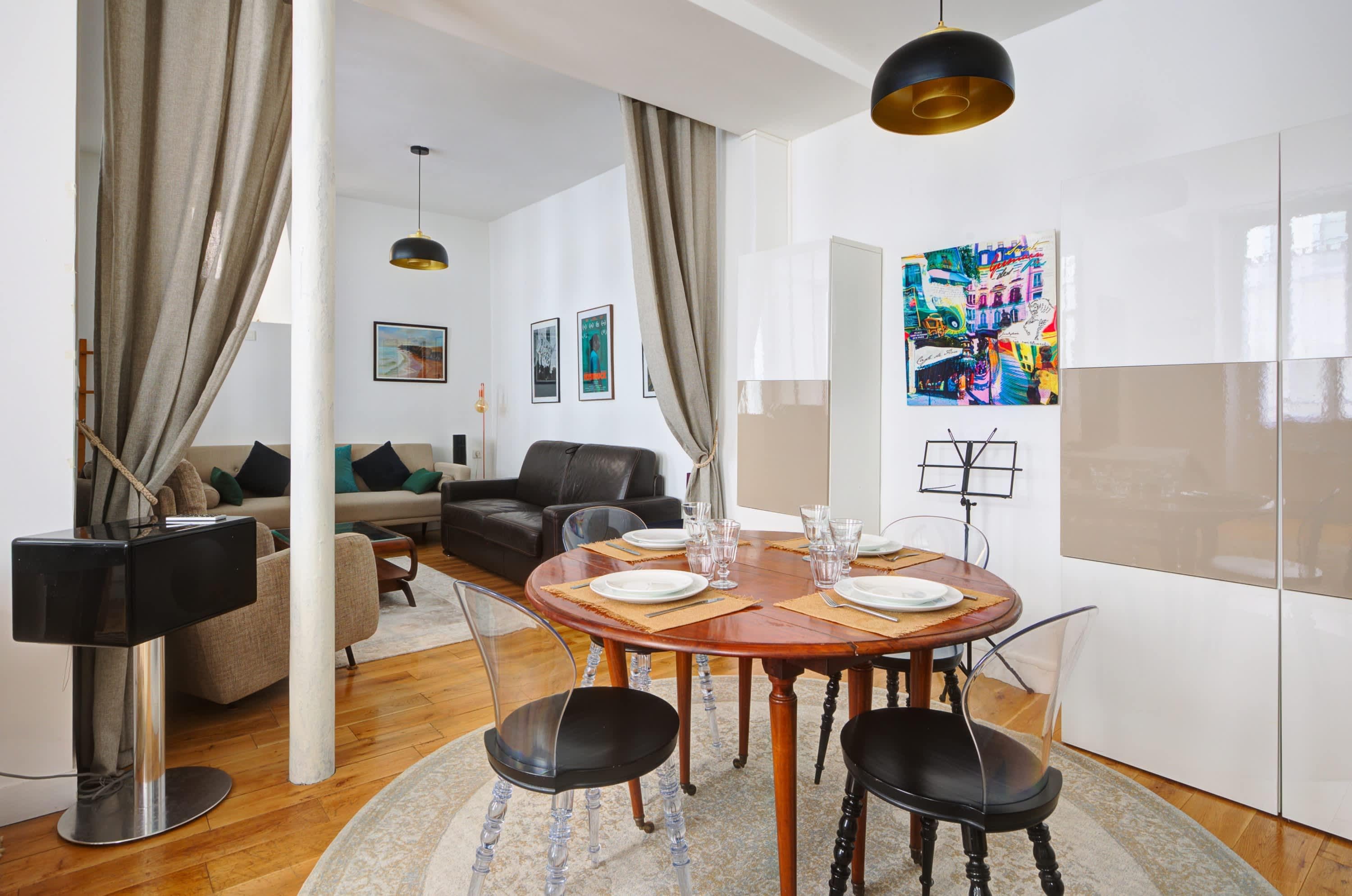 A modern living room with a round wooden dining table set for four, surrounded by black chairs. There is a sofa and a coffee table in the background.