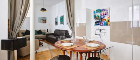 Un salon moderne avec une table de salle à manger ronde en bois, disposée pour quatre personnes, entourée de chaises noires. On aperçoit un canapé et une table basse au fond de la pièce.