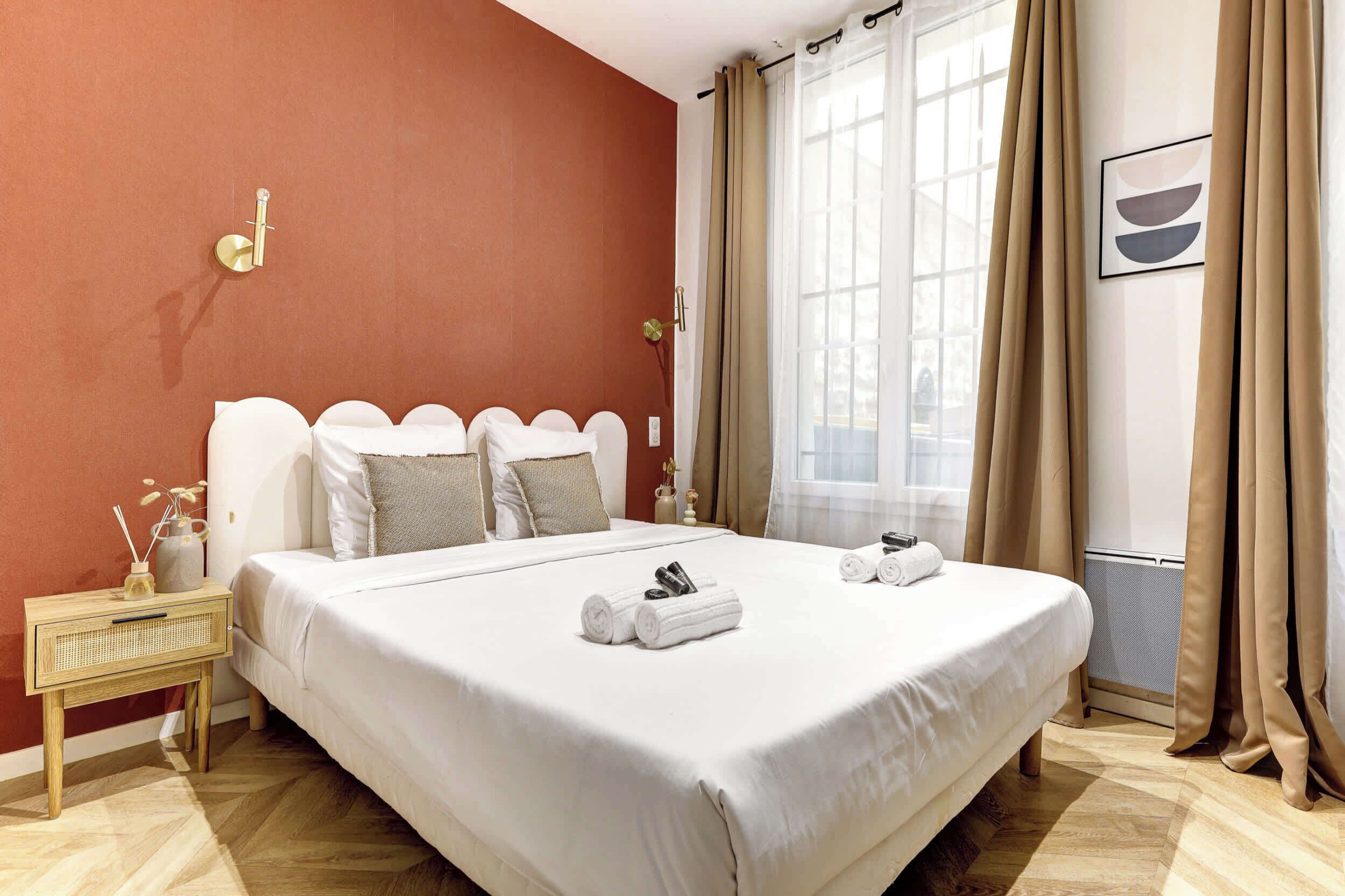 A neatly made bed with white linens, a bedside table with a vase and a lamp, and a window with beige curtains.