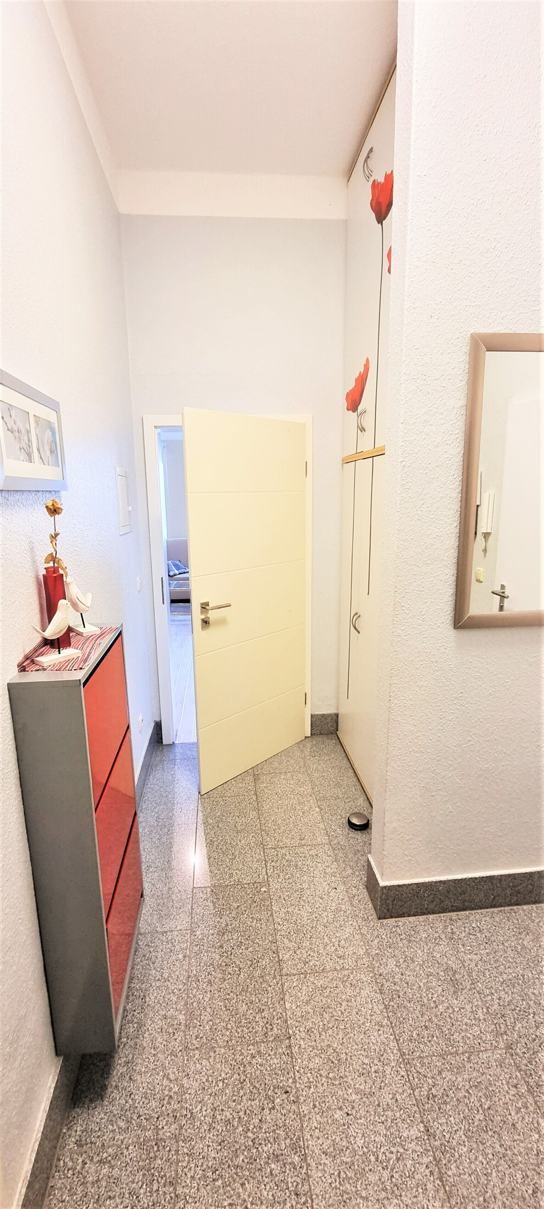 Un pasillo con suelo de baldosas, una puerta cerrada, un aparador con un florero y un espejo montado en la pared.