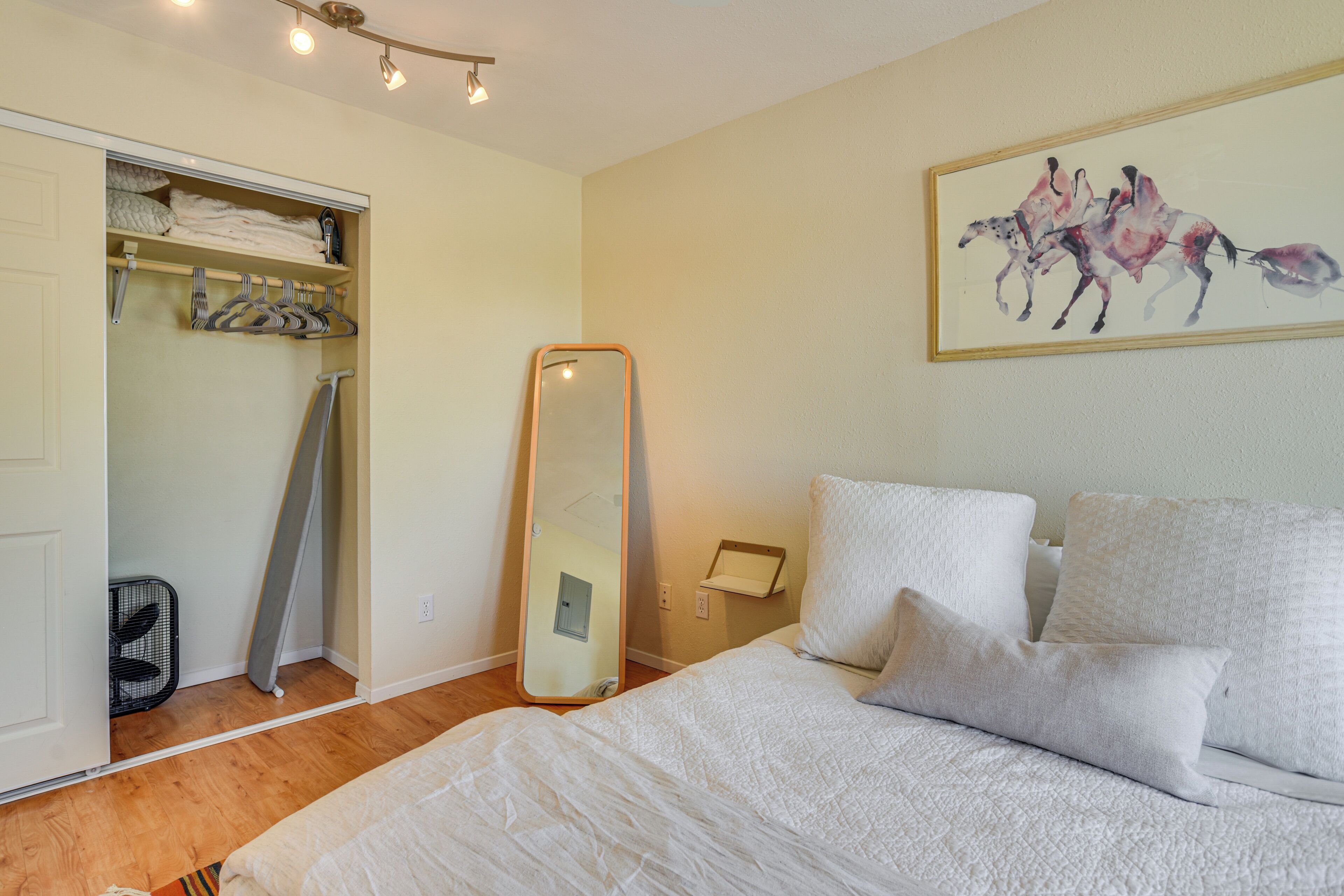 Una camera da letto con un letto, cuscini, un grande specchio, un armadio con abiti appesi e un quadro sulla parete.