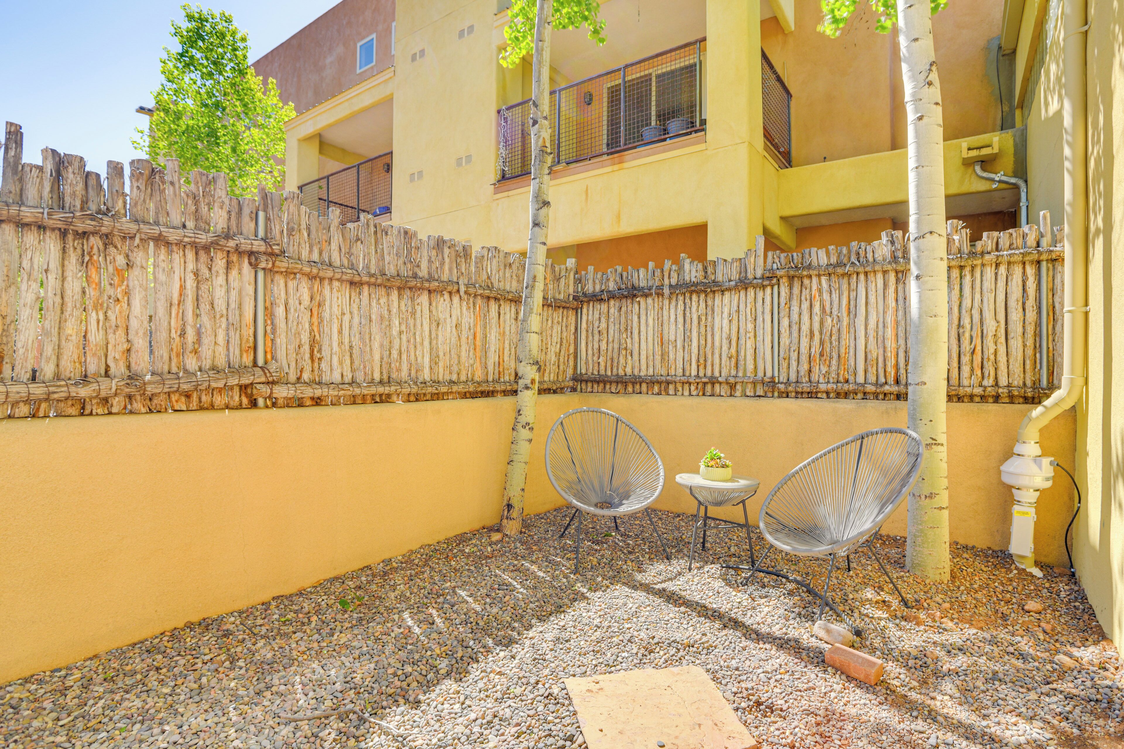 A courtyard with a wooden fence, two metal chairs, a small table, and a tree.