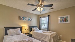A bedroom with two beds, a ceiling fan, a window with blinds, a painting, and a 'RELAX' sign.