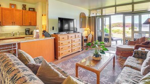 A spacious living area with a sofa, a coffee table, and a television. There is a kitchen area with wooden cabinets and a coffee machine. - Oceanfront Condo w/ Pool Access on Molokai! (Maunaloa)