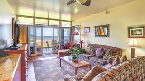 A spacious living room with a large sofa, a coffee table, and a television. There is a sliding glass door leading to a balcony with a view of the ocean. - Oceanfront Condo w/ Pool Access on Molokai! (Maunaloa)