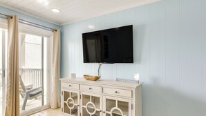 A room with a TV mounted on the wall, a wooden cabinet with drawers, a glass table, and a sliding glass door leading to an outdoor area. - 1 Mi to Barefoot Landing: Condo w/ Pool Access! (North Myrtle Beach)
