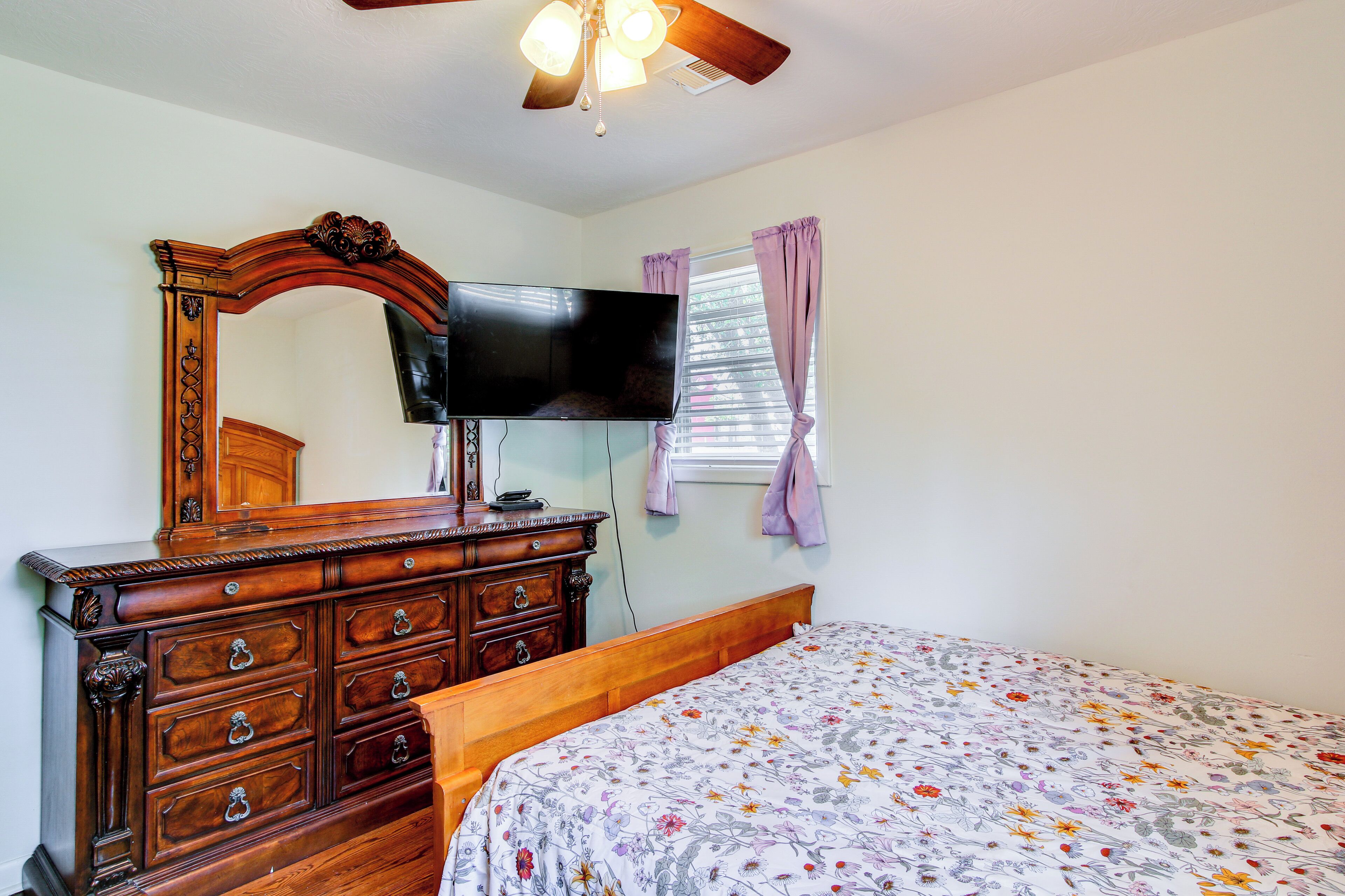 Une chambre avec une commode en bois, un téléviseur à écran plat fixé au-dessus, un lit recouvert d’une couvre-lit à motifs floraux et une fenêtre ornée de rideaux violets.