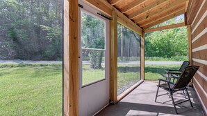 Eine überdachte Veranda mit großen Fenstern, einem Sessel und Blick auf eine Rasenfläche und Bäume.