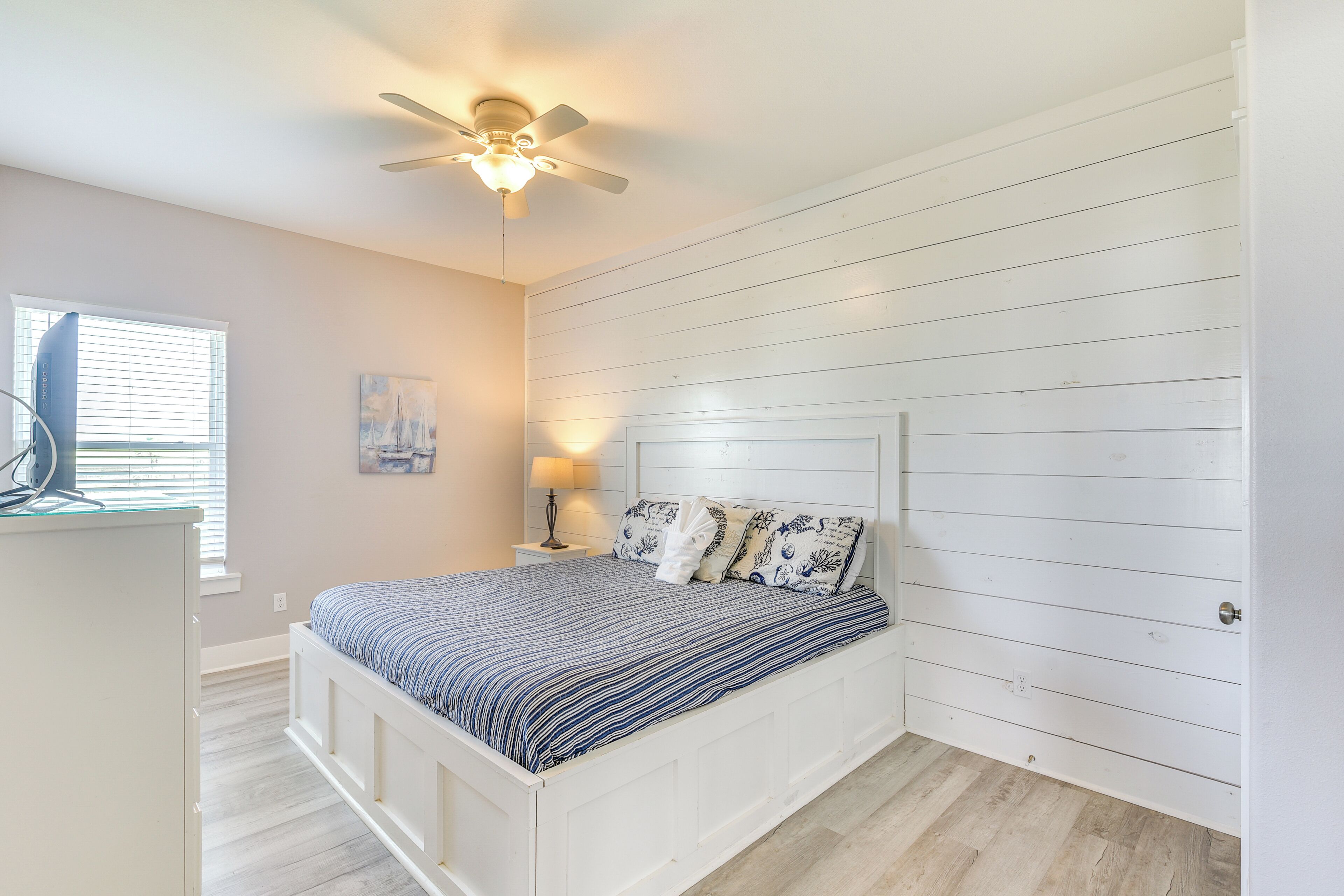 A bedroom with a bed, a ceiling fan, a window with blinds, and a nightstand with a lamp.