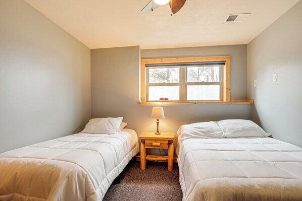 A bedroom with two single beds, a small wooden nightstand with a lamp, and a window with a view of trees.
