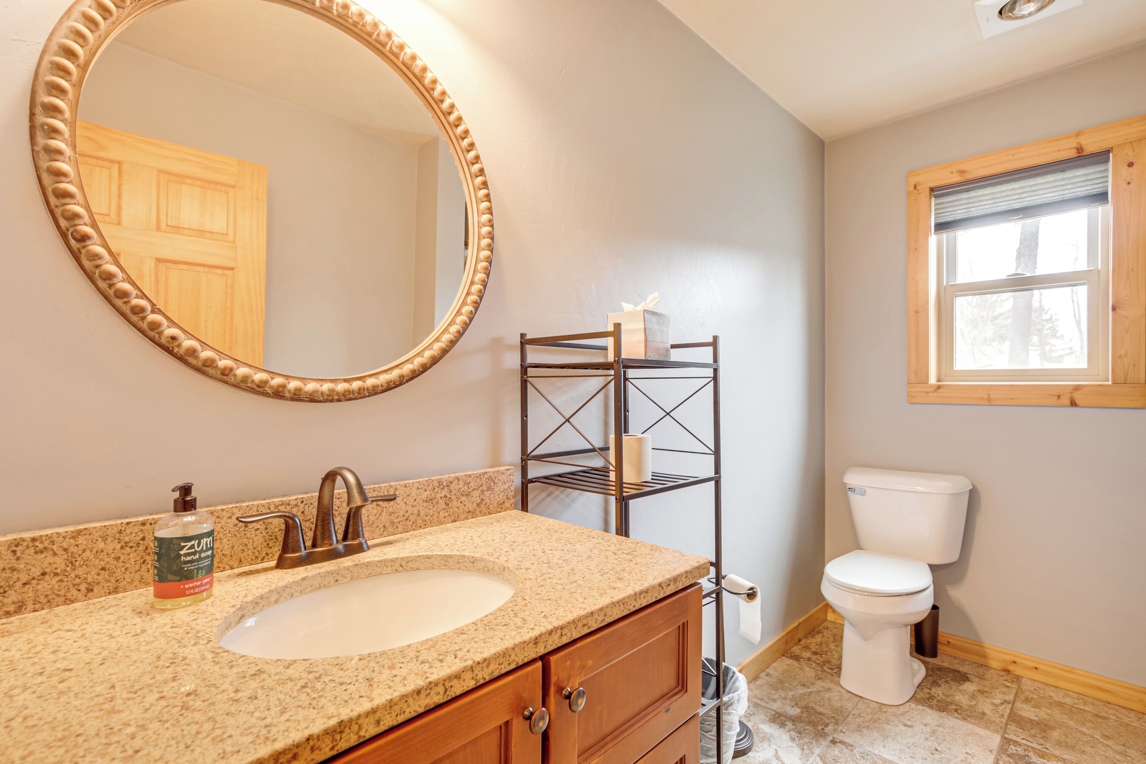 A bathroom with a granite countertop sink, a wooden vanity, a freestanding toilet, a towel rack, and a window with a wooden frame.
