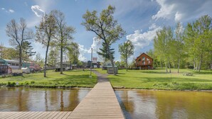 A wooden dock leading to a calm water body, surrounded by green grass and trees, with a building in the background. - Pet Friendly Lake House: Waterfront w/ Boat Dock (Eagle River)