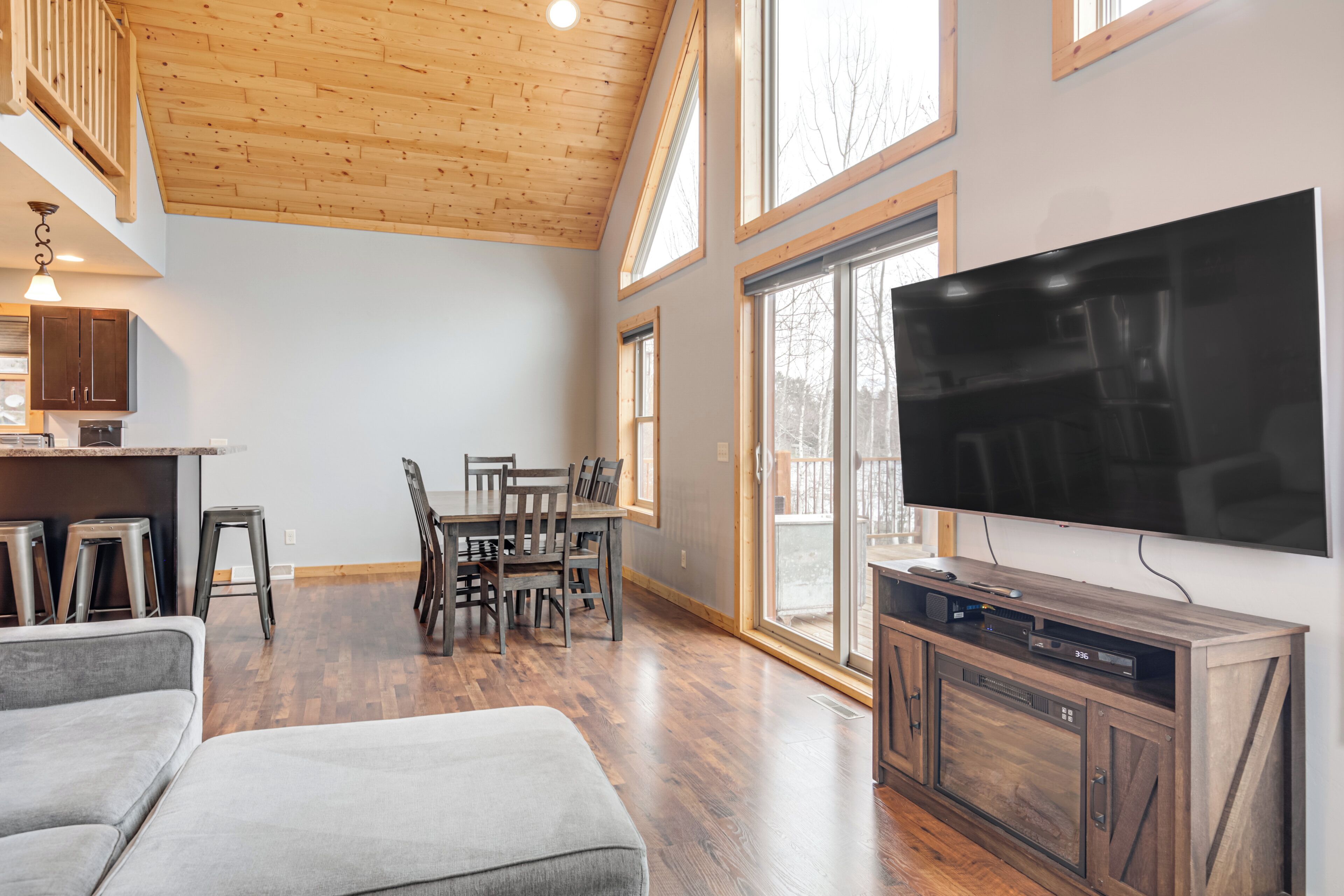 A spacious living area with a wooden ceiling, a flat-screen TV mounted on the wall, a dining table with chairs, and a gray sofa.
