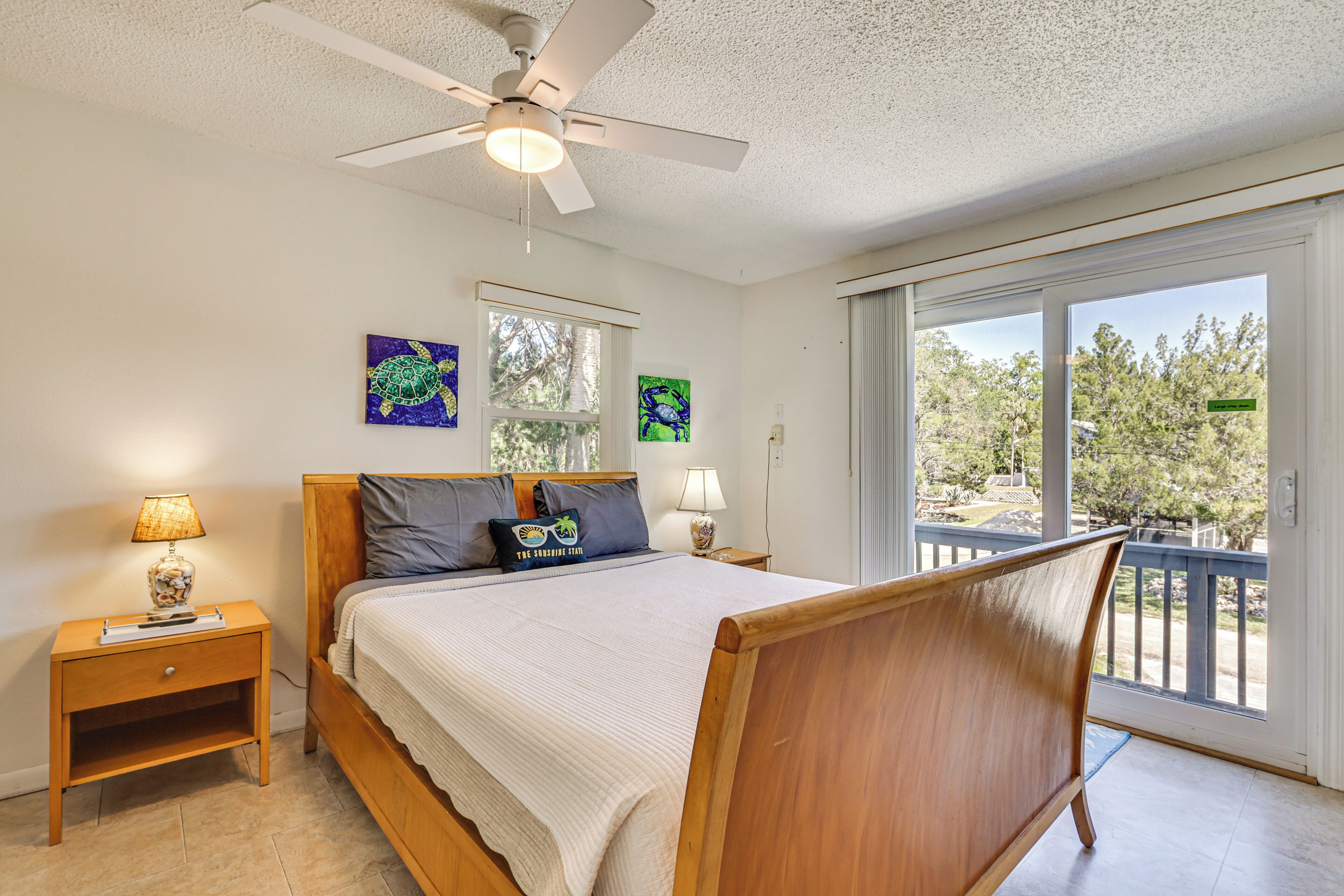 Quarto com cama, cabeceira de madeira, duas mesas de cabeceira com abajures, um ventilador de teto e uma janela grande com vista para árvores.