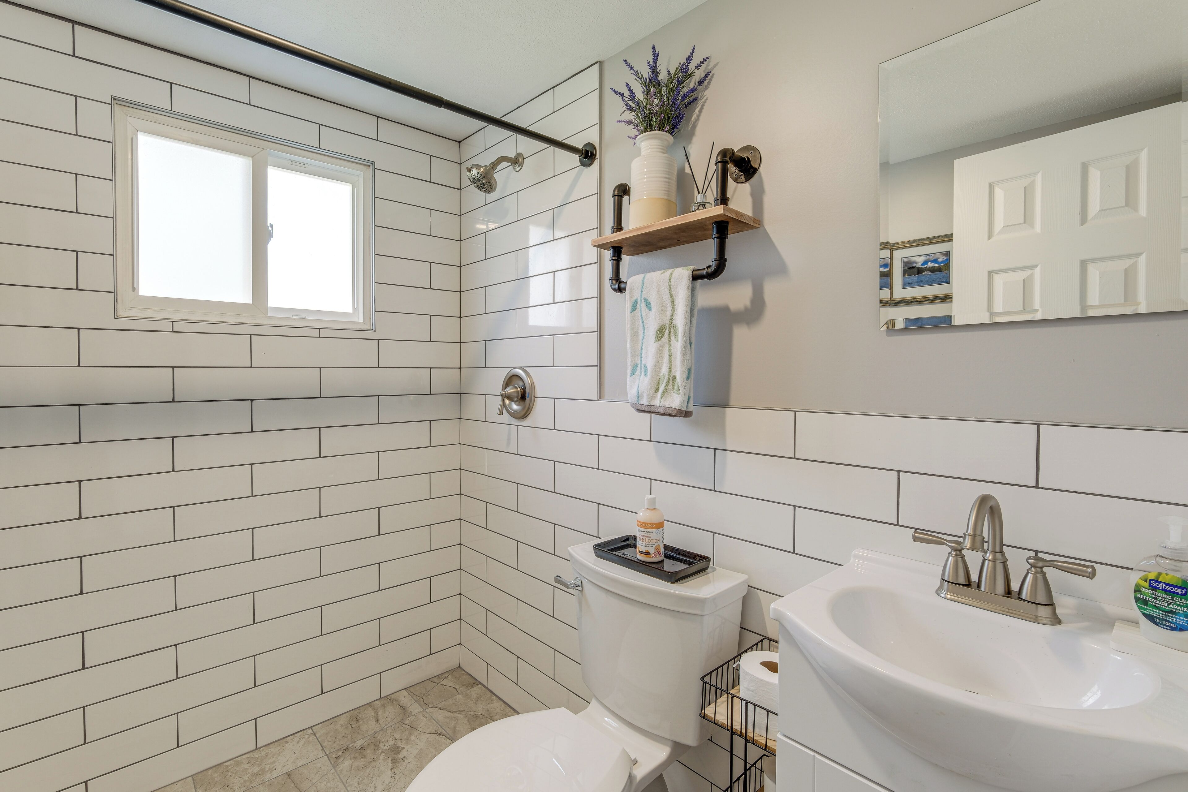 Une salle de bain avec des carreaux de céramique blancs rectangulaires, des toilettes, un lavabo avec un robinet, une fenêtre et une étagère avec une plante et une serviette.