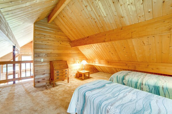 A cozy attic bedroom with wooden walls and a sloped ceiling. There is a bed with a blue and white striped bedspread, a wooden dresser, and a small table with a lamp. - Hot Tub, 1 Mi to Beach: 12-acre Fort Bragg Cabin! (Fort Bragg)