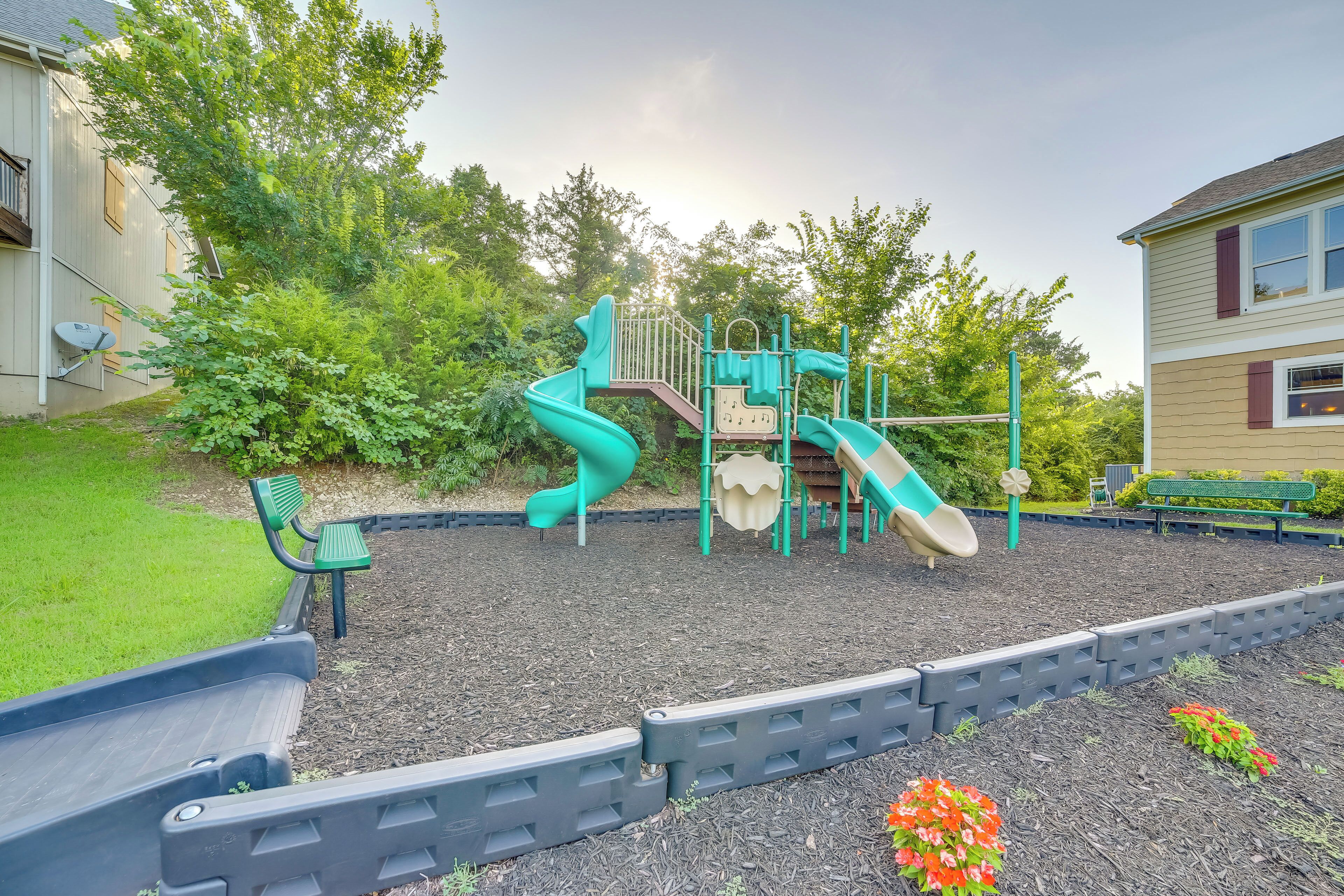 A playground with slides and climbing structures, surrounded by a garden with flowers and a bench.
