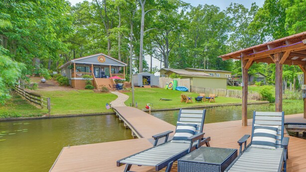 A deck with lounge chairs and a table overlooking a pond, surrounded by a wooden deck and a small building. - Private Dock + Porch: Lakefront Retreat in Cobb! (Cobb)