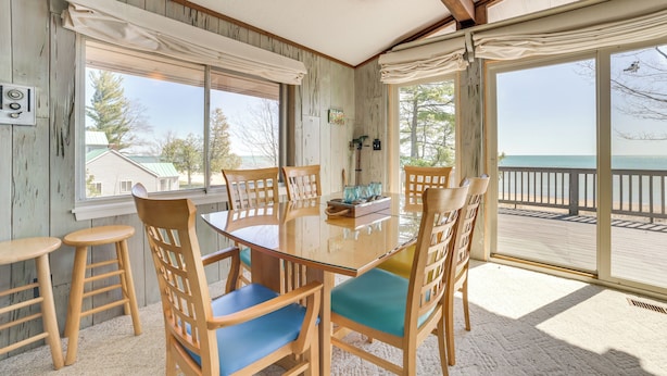 A dining area with a glass table and wooden chairs, large windows with curtains, and a view of the beach. - Private Beach: Waterfront Home on Lake Huron! (Greenbush)